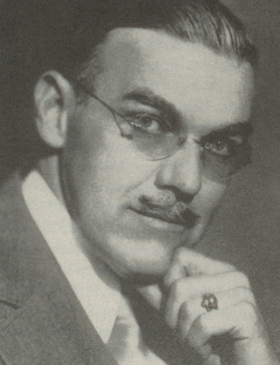A man in glasses is wearing a suit, has his head tilted down and to the side, and is looking at the camera.